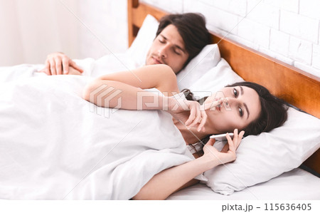 A woman is lying in bed next to a sleeping man, holding a phone to her ear while whispering into it with her finger on her lips. The man appears to be asleep, unaware of secretive phone call. 115636405