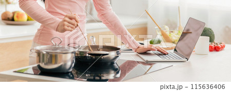 A woman in a pink shirt stirs food in a frying pan while referencing her laptop in a bright, modern kitchen filled with fresh ingredients, cropped 115636406