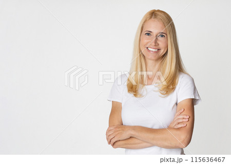 A smiling blonde woman with long hair is wearing a white t-shirt and has her arms crossed in front of her. She is standing in front of a white background, copy space A smiling blonde woman with long hair is wearing a white t-shirt and has her arms crossed in front of her. She is standing in front of a white background, copy space 115636467