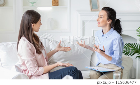 Two women are seated on a comfortable white sofa having an engaging conversation. One woman appears to be a therapist holding a clipboard, while the other, dressed casually, seems to be the client. 115636477