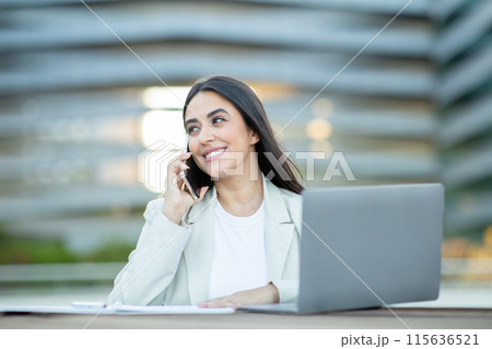 A young woman in a white blazer smiles while talking on her phone, with a laptop in front of her. She is sitting outside in front of a modern building. 115636521