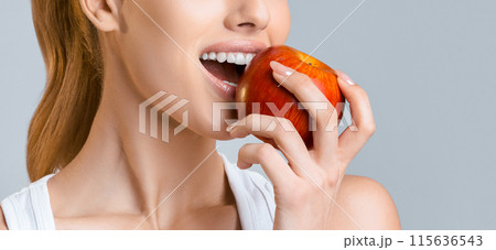 A close-up shot of a woman taking a bite out of a red apple. She is wearing a white tank top and her hair is pulled back in a ponytail. The photo is taken against a light grey background. A close-up shot of a woman taking a bite out of a red apple. She is wearing a white tank top and her hair is pulled back in a ponytail. The photo is taken against a light grey background. 115636543