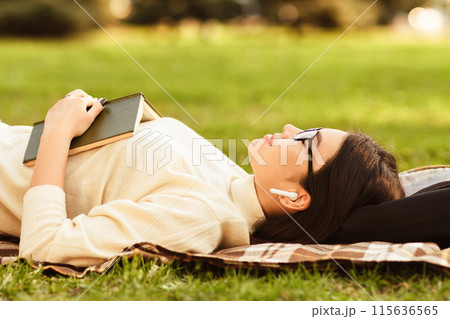 Asian woman is lying on a blanket in a park during a sunny afternoon, holding a book and wearing earbuds. She appears to be enjoying a peaceful moment, possibly listening to music Asian woman is lying on a blanket in a park during a sunny afternoon, holding a book and wearing earbuds. She appears to be enjoying a peaceful moment, possibly listening to music 115636565