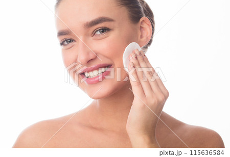 Closeup of woman is smiling as she applies facial cleanser to her skin. Her expression reflects satisfaction and relaxation while she takes care of her skincare routine. 115636584