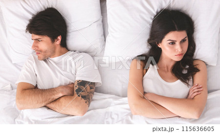 A couple is lying in bed with their arms crossed, looking away from each other. The woman is looking to the right with a serious expression, and the man is looking down with a furrowed brow. 115636605