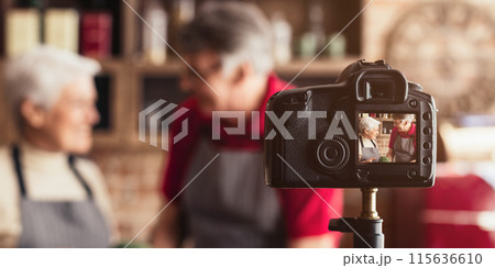 A happy senior couple, wearing aprons, stand in a home kitchen and film a cooking video. The couple smiles as they look at the camera, which is mounted on a tripod. 115636610