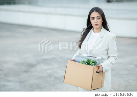 A young woman, dressed in a white blazer and a white shirt, walks away from a building, carrying a cardboard box filled with personal items. She has a serious expression on her face. A young woman, dressed in a white blazer and a white shirt, walks away from a building, carrying a cardboard box filled with personal items. She has a serious expression on her face. 115636636