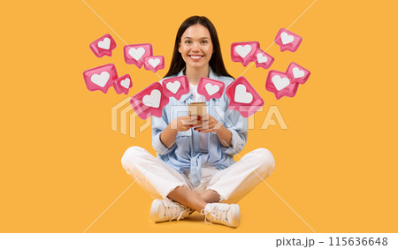 Cheerful young european woman in casual clothes sits cross-legged holding smartphone, chatting with guys on dating app, smiling at camera against vibrant yellow background 115636648