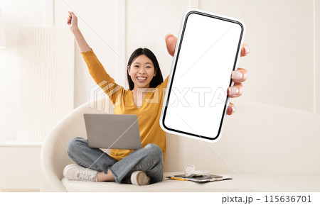 A cheerful young asian woman, dressed in a casual yellow top and jeans, sits cross-legged on a cozy white sofa with her laptop, enthusiastically presenting a smartphone with a blank screen 115636701