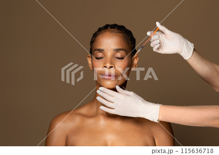 Cosmetic beauty injections concept. Attractive black middle aged woman getting facial injection in cheekbones zone, standing over brown studio background, free space Cosmetic beauty injections concept. Attractive black middle aged woman getting facial injection in cheekbones zone, standing over brown studio background, free space 115636718