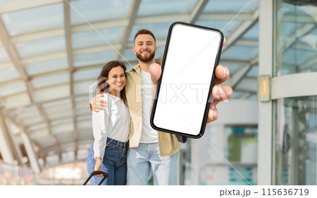 A cheerful man and woman stand together in an airport terminal, with the man extending his arm towards the camera, showing a smartphone with a blank screen 115636719