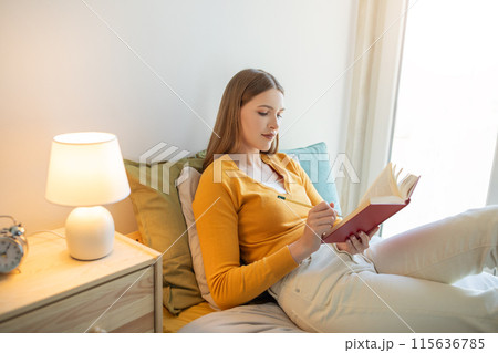 A girl is seated on a bed, engrossed in reading a book. She is dressed casually and appears focused on the content of the book. 115636785