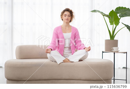 Calm young woman sitting on couch, meditating with closed eyes at home, peaceful beautiful female practicing yoga in lotus pose, relaxing on sofa, coping with inner stress, copy space 115636799