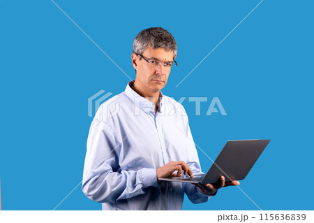 Professional IT specialist. Mature businessman holding laptop computer typing and working online, frowning with serious face on blue background. Man browsing internet, web business career concept 115636839