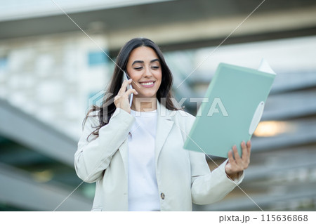 A young woman in a white blazer smiles as she talks on her mobile phone while holding a blue folder. She is outdoors in an urban setting, likely on a business call. 115636868