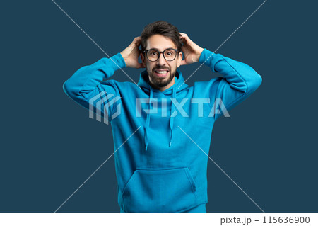 A man wearing a blue hoodie is standing with his hands pressed against his ears, appearing to be in distress or experiencing loud noise. He looks tense and focused on the sounds he is hearing. A man wearing a blue hoodie is standing with his hands pressed against his ears, appearing to be in distress or experiencing loud noise. He looks tense and focused on the sounds he is hearing. 115636900