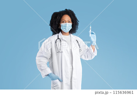 Confident serious young black lady doctor in white coat, protective mask and gloves shows syringe, isolated on blue studio background. Disease treatment, vaccination, medicine and health care 115636961