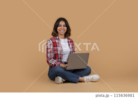 A young middle eastern woman is sitting on a beige floor. She is holding a laptop computer and smiling at the camera. 115636981