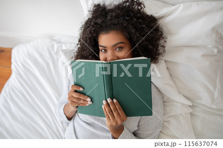 Hispanic woman is laying comfortably in bed, engrossed in a book she is reading. The room is softly lit, top view 115637074
