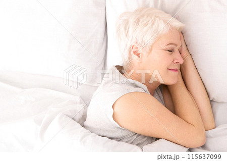 A woman is lying down in bed, covered by crisp white sheets. She appears relaxed and comfortable, with her eyes closed, possibly resting or sleeping, view above 115637099