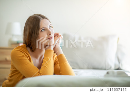 A girl is seated on a neatly made bed, with one hand resting gently on her chin. She appears deep in thought, her posture reflective and pensive. A girl is seated on a neatly made bed, with one hand resting gently on her chin. She appears deep in thought, her posture reflective and pensive. 115637155