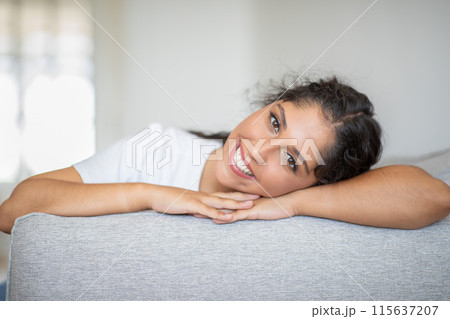 A woman with curly dark hair rests on a gray couch, her head propped on her arms. She smiles broadly and looks directly at the camera. She is wearing a simple, white t-shirt. A woman with curly dark hair rests on a gray couch, her head propped on her arms. She smiles broadly and looks directly at the camera. She is wearing a simple, white t-shirt. 115637207