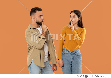 A young man and woman stand facing each other, both holding a finger to their lips, signaling for silence against a solid orange background. They seem to be engaged in a quiet conversation or secret. 115637280