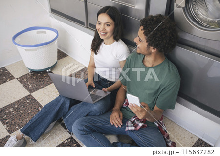 Leisure At Laundry. Cheerful Students Couple With Laptop And Smartphone Sitting In Washing Room With Industrial Washer Machines, Waiting For Clean Clothes At Laundrette Indoors Leisure At Laundry. Cheerful Students Couple With Laptop And Smartphone Sitting In Washing Room With Industrial Washer Machines, Waiting For Clean Clothes At Laundrette Indoors 115637282