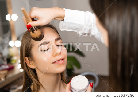 Make up artist applying powder with a brush to a blonde woman at beauty salon 115637286