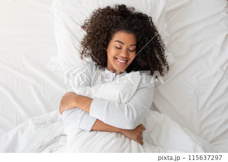 A cheerful young Hispanic woman with curly hair is hugging a white pillow while lying in a cozy, sunlit bed. Her eyes are gently closed, and her expression is one of comfort and happiness A cheerful young Hispanic woman with curly hair is hugging a white pillow while lying in a cozy, sunlit bed. Her eyes are gently closed, and her expression is one of comfort and happiness 115637297