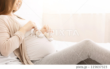 A pregnant woman is seen resting comfortably on bed, holding a pair of baby shoes near her belly. She appears content and serene, enjoying a quiet moment, cropped 115637323