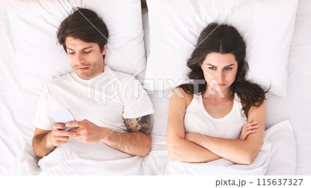 A young woman with long dark hair lies in bed with her arms crossed, looking upset. Her male partner is lying next to her, using his phone. The scene depicts a potential relationship issue A young woman with long dark hair lies in bed with her arms crossed, looking upset. Her male partner is lying next to her, using his phone. The scene depicts a potential relationship issue 115637327