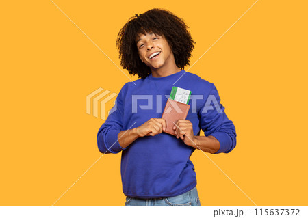 A joyful young man in a blue sweater stands against a vibrant yellow background, holding his passport and flight tickets, ready for his upcoming trip. 115637372