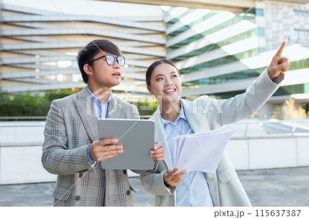 Two young Asian business professionals stand together outside an office building. heir attire and gadgets suggest a work-related interaction during daylight, using digital tablet Two young Asian business professionals stand together outside an office building. heir attire and gadgets suggest a work-related interaction during daylight, using digital tablet 115637387