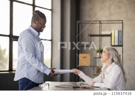 Job Interview Concept. HR Manager Lady Giving Papers To African American Business Man Making Career Offer To Applicant At Modern Office Interior. Corporate Meeting And Employment. Side View Shot 115637416