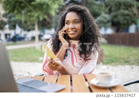 Smiling black woman holding credit card and talking on cellphone outdoors, happy young african american lady sitting at table in cafe, buying online on laptop, enjoying easy payments, closeup Smiling black woman holding credit card and talking on cellphone outdoors, happy young african american lady sitting at table in cafe, buying online on laptop, enjoying easy payments, closeup 115637417