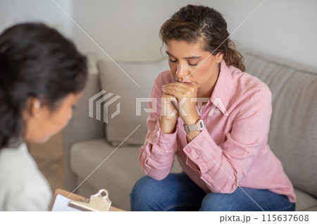 A young woman, wearing a pink shirt and jeans, sits on a beige couch, looking down with her hands clasped together in her lap. She appears to be in a counseling session 115637608