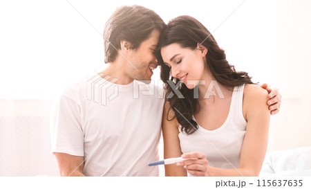 A smiling couple is sitting on a bed at home, with the woman holding a positive pregnancy test. The man has his arm around the woman and they are looking at each other with love and happiness. A smiling couple is sitting on a bed at home, with the woman holding a positive pregnancy test. The man has his arm around the woman and they are looking at each other with love and happiness. 115637635