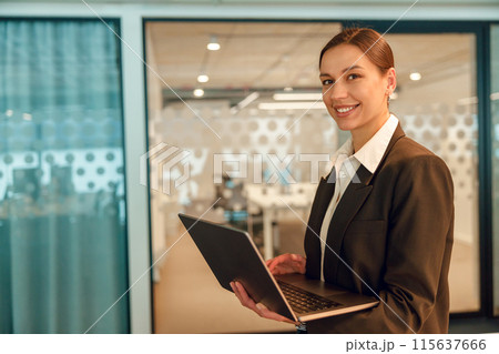 A businesswoman holding a laptop in a modern office, looking professional and successful 115637666