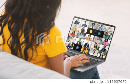 A young black woman is engaged in a virtual team meeting while working from her home office. She is seated, looking at a laptop screen displaying numerous colleagues via video call A young black woman is engaged in a virtual team meeting while working from her home office. She is seated, looking at a laptop screen displaying numerous colleagues via video call 115637672