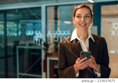 A businesswoman is smiling as she uses a smartphone in a contemporary office environment 115637675