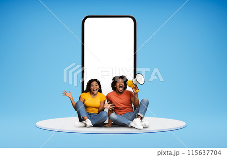 Exciting online offer, deal. Thrilled millennial black couple hipsters posing by huge phone with white blank screen, sitting on platform over blue background, making announcement via loudspeaker 115637704