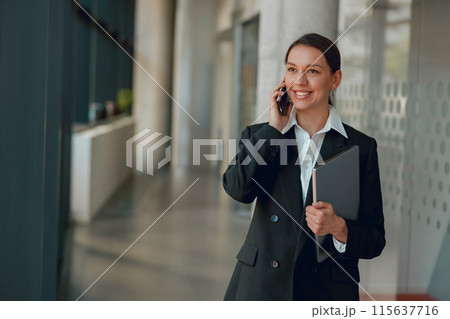 A professional woman in office is talking on phone while holding a folder and smiling 115637716