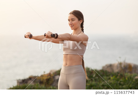 Positive millennial caucasian lady athlete in sportswear doing arm exercises with dumbbells, enjoy workout at sea beach. Fitness outdoor, sports, body care alone and active lifestyle Positive millennial caucasian lady athlete in sportswear doing arm exercises with dumbbells, enjoy workout at sea beach. Fitness outdoor, sports, body care alone and active lifestyle 115637743
