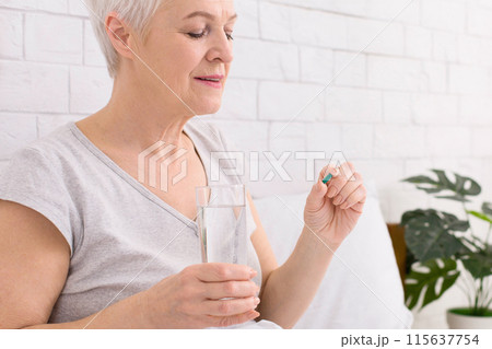 Senior woman with short grey hair is gently holding a pill in one hand and a clear glass of water in the other. She appears calm and focused, preparing to take her medication after wake up 115637754