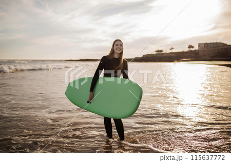 Sport Activities. Portrait Of Cheerful Young Surfer Woman Posing With Surfboard, Smiling Millennial Female Wearing Wetsuit Surfing On The Beach On Sunset, Enjoying Water Sports And Summer Vacation 115637772