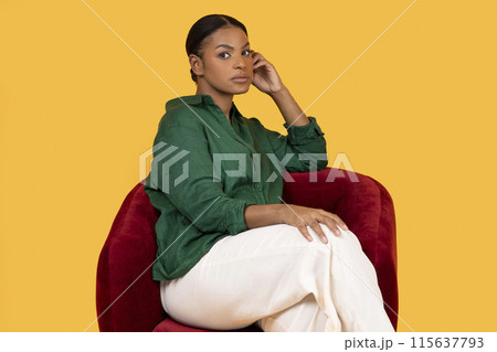 African American Woman In Casual Green Shirt Looking At Camera Sitting In Red Armchair Over Yellow Background. Studio Shot Calm Lady Thinking And Posing For Advertisement Banner 115637793