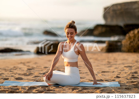 Young european lady athlete in sportswear sitting in lotus position practicing yoga, enjoying stretching body exercises on sea beach. Fit outdoor, sport and weight loss 115637823
