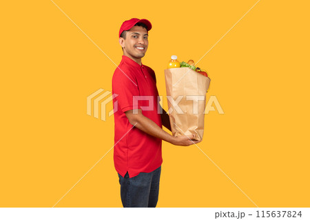 Delivery man standing over yellow studio background, holding a paper bag filled with various food items. He is looking directly at the camera Delivery man standing over yellow studio background, holding a paper bag filled with various food items. He is looking directly at the camera 115637824