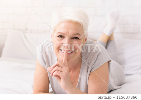 A cheerful senior woman with short white hair lies on her stomach on a bed, her eyes beaming with joy. She places her finger on her lips, suggesting a playful secret or a quiet moment A cheerful senior woman with short white hair lies on her stomach on a bed, her eyes beaming with joy. She places her finger on her lips, suggesting a playful secret or a quiet moment 115637860